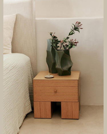 Mesita de noche de chapa de roble con un cajón de Ferm Living: mesita de un cajón junto a cama clara, con jarrón verde ondulado y ramas.