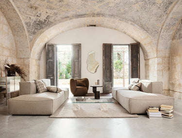 Módulo de Ferm Living: sala de arco de piedra con dos composiciones modulares claras enfrentadas, sillón marrón y mesa de vidrio; foto horizontal con bandas lisas superior e inferior.