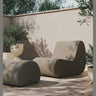 Módulo de sofá de exterior en bouclé arena de Ferm Living: terraza: módulo Rouli junto a puf, pavimento claro y vegetación; sombra de hojas sobre tapicería arena oscura.