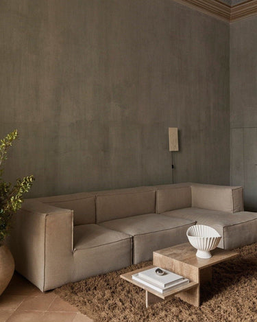 Módulo de sofá profundo de lino con brazo izquierdo de Ferm Living: ambiente de sofá modular de varios asientos frente a pared gris, mesa baja pétrea y jarrón blanco.