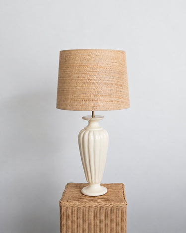Pantalla de lámpara de Mahe Homeware sobre una base blanca y un pedestal de ratán.