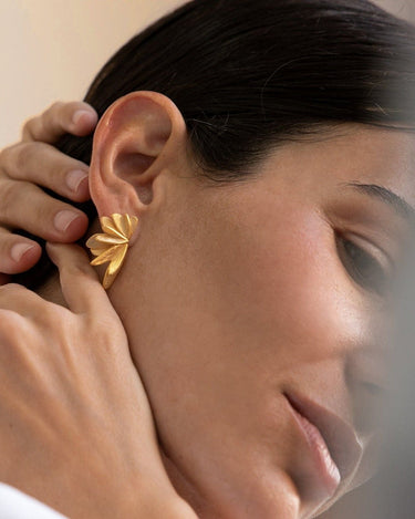 Par de pendientes dorados de formas desplegadas de Joidart: pendiente dorado de elementos alargados desplegados como abanico puesto en el lóbulo; mano junto al cabello.
