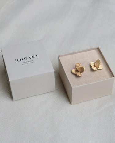 Dos pendientes dorados pequeños de flor en caja clara con tapa Joidart.