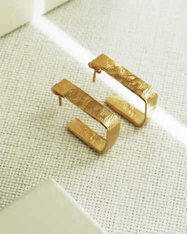 Dos aros rectangulares dorados anchos vistos oblicuamente sobre tejido claro.