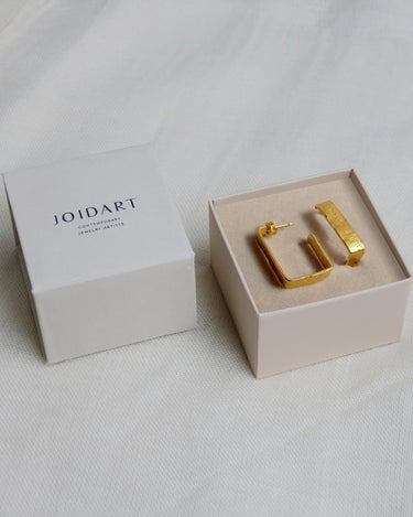 Par de aros rectangulares dorados anchos en caja clara con tapa Joidart al lado.