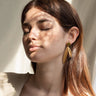 Par de pendientes largos de dos hojas doradas de Joidart: pendiente largo con círculo verde y dos hojas doradas puesto; modelo bajo luz y sombras vegetales.