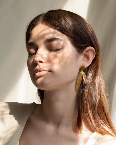 Par de pendientes largos de dos hojas doradas de Joidart: pendiente largo con círculo verde y dos hojas doradas puesto; modelo bajo luz y sombras vegetales.