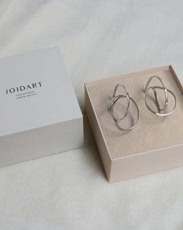 Dos pendientes plateados grandes en caja clara con tapa Joidart al lado.