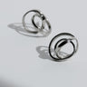 Par de pendientes de plata pequeños entrelazados de Joidart: dos pendientes pequeños plateados de cintas entrelazadas sobre fondo gris con sombras.