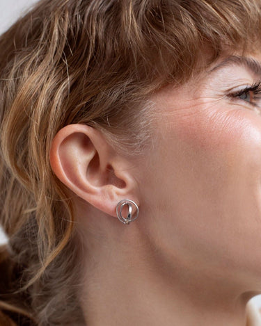 Uno de los pendientes pequeños plateados Embolic de Joidart puesto en el lóbulo, con el rostro de perfil y el cabello recogido.