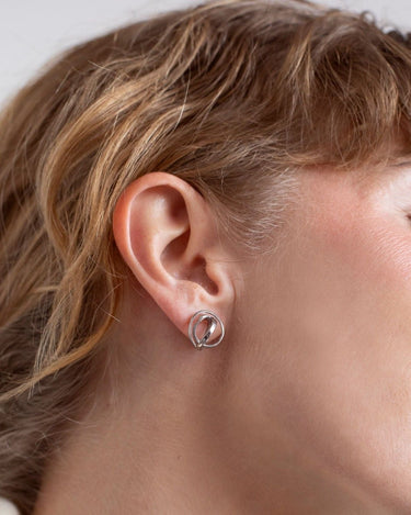 Par de pendientes de Joidart: pendiente pequeño plateado puesto en el lóbulo, visto de perfil; cabello recogido.