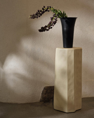 Pedestal de hormigón blanco roto de Ferm Living con jarrón negro y flores encima.