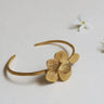 Pulsera dorada abierta con flor y hoja de Joidart: pulsera dorada abierta con flor estriada y hoja, sobre fondo claro con pequeñas flores de atrezzo.