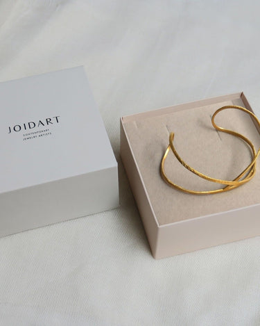 Pulsera dorada abierta en caja clara, con tapa de Joidart a un lado.