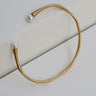 Pulsera dorada sinuosa con perlas de vidrio de Joidart: pulsera dorada abierta y sinuosa con dos extremos blancos nacarados, sobre planos claros.