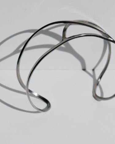 Pulsera de Joidart: pulsera plateada abierta con sombra sobre fondo gris; parte del extremo derecho queda fuera del encuadre.