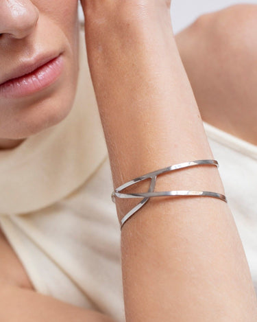 Pulsera de plata abierta de cintas entrelazadas de Joidart: pulsera plateada de cintas entrelazadas puesta en muñeca junto al rostro de una modelo.