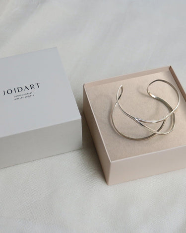 Una de las pulseras plateadas Embolic dentro de una caja clara, con la tapa de Joidart a la izquierda.