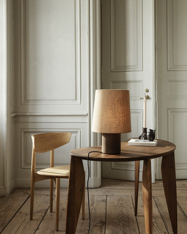 Silla de Ferm Living: silla junto a mesa redonda oscura y lámpara de sobremesa, ante pared y puerta con molduras.