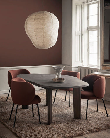 Silla de Ferm Living: varias sillas tapizadas alrededor de mesa redonda oscura, bajo lámpara de papel.