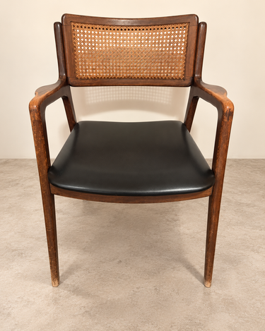 Silla vintage de madera y rejilla con asiento negro, vista frontal sobre suelo de cemento topo claro