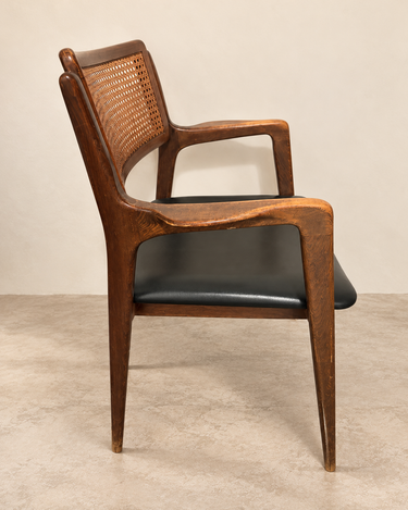 Silla vintage de madera y rejilla con asiento negro, vista lateral sobre suelo de cemento topo claro