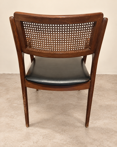 Silla vintage de madera y rejilla con asiento negro, vista trasera sobre suelo de cemento topo claro
