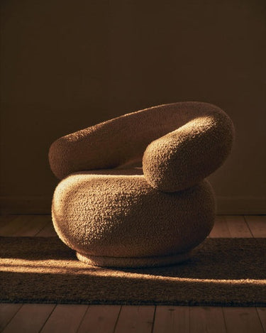 Sillón de bouclé con respaldo curvo de Ferm Living, en marrón y en un interior con luz cálida.