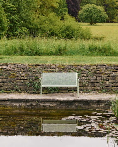 Sofá de jardín de dos plazas de acero perforado de Ferm Living: sofá verde de dos plazas junto a estanque, reflejado en el agua frente a muro de piedra.