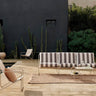 Sofá de jardín de tres plazas con cojines a rayas de Ferm Living: sofá de tres plazas con rayas claras y oscuras en terraza, mesa ligera y varios asientos; bandas de fondo liso arriba y abajo.