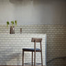 Taburete de bar de roble oscuro y tapizado gris de MUUBS junto a una pared de azulejos
