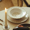Vajilla de gres con hojas pintadas en beige de Leandro Cano: platos blanco y beige sobre mesa con silla de rejilla y vela en primer plano.