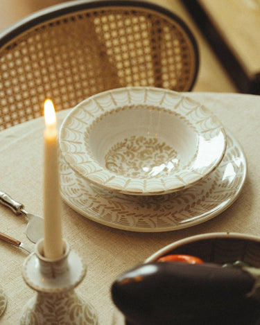 Vajilla de gres con hojas pintadas en beige de Leandro Cano: platos blanco y beige sobre mesa con silla de rejilla y vela en primer plano.