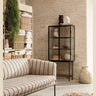 Vitrina alta de acero negro y cristal estriado de Ferm Living: vitrina alta negra al fondo de un salón con sofá a rayas, estantería y plantas.
