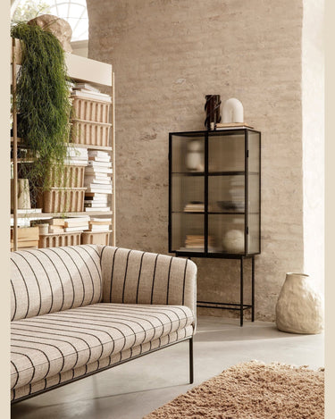 Vitrina alta de acero negro y cristal estriado de Ferm Living: vitrina alta negra al fondo de un salón con sofá a rayas, estantería y plantas.