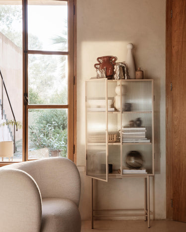 Vitrina de Ferm Living junto a una ventana, con una puerta abierta y objetos en los estantes.