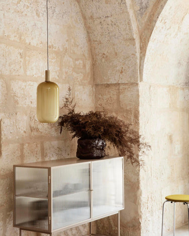 Vitrina baja de acero y cristal estriado de Ferm Living, acero beige: vitrina beige junto a un muro de piedra con jarrón de ramas.